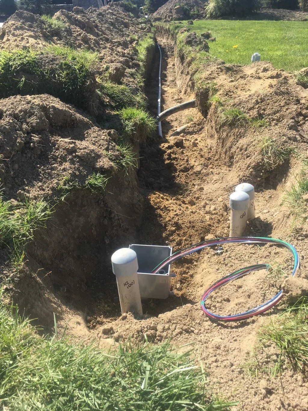 A trench in the ground with gray pipes and colored wires, outdoors on a grassy lawn.