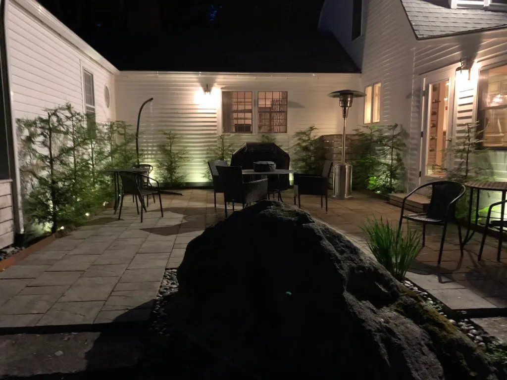 Nighttime view of a backyard patio with a grill, chairs, and surrounding greenery, illuminated by lights.