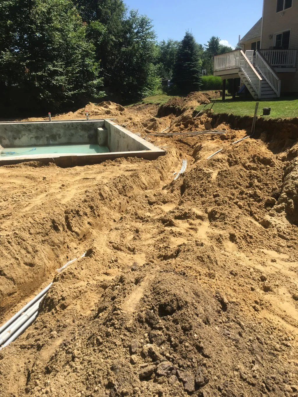 Pool construction site; dirt excavated around a rectangular concrete pool shell, piping visible. Backyard setting.