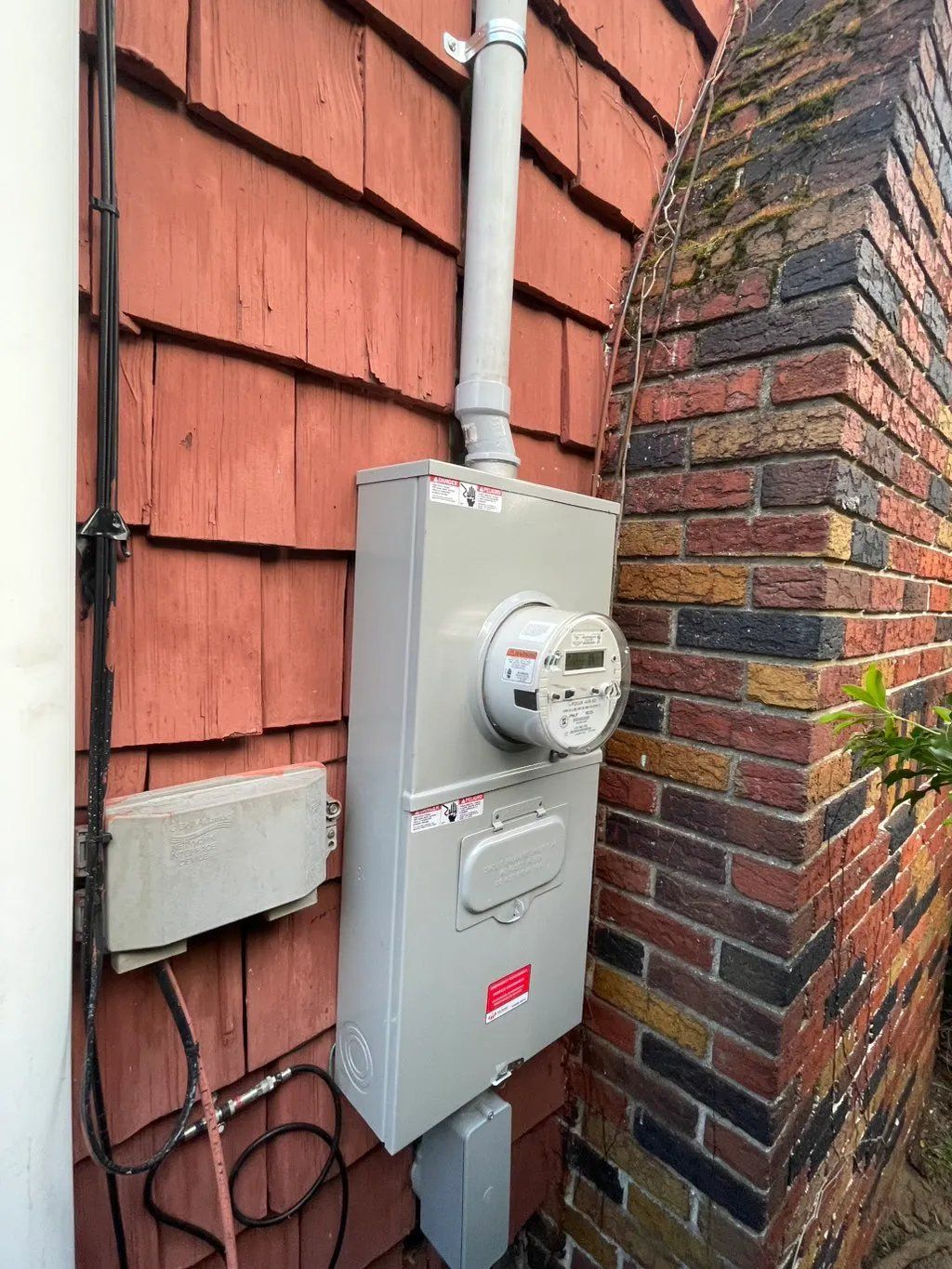 Gray electrical meter box mounted on a red shingled wall next to a brick chimney.