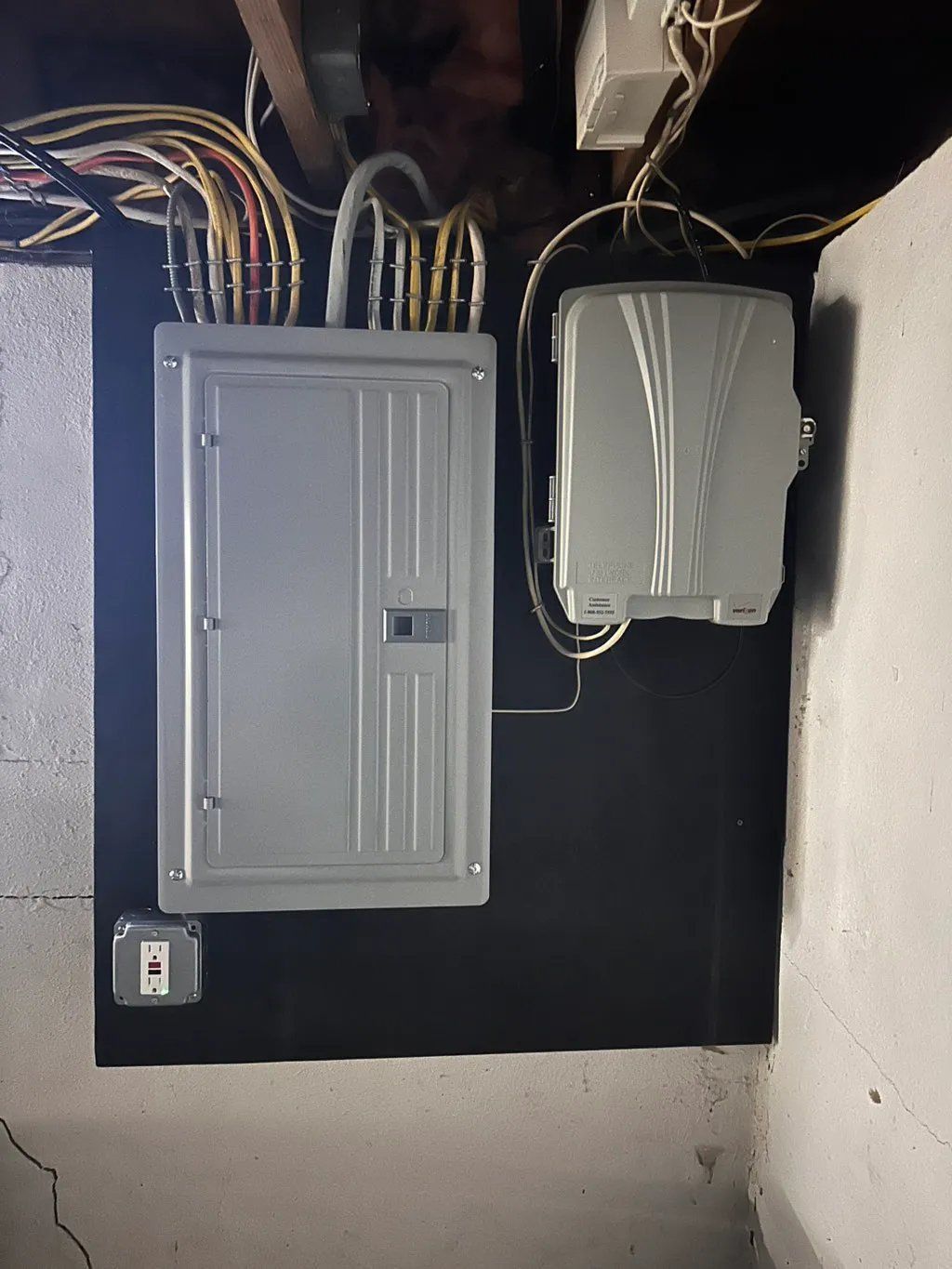 Electrical panel and telecommunications box mounted on a black surface in a basement corner.