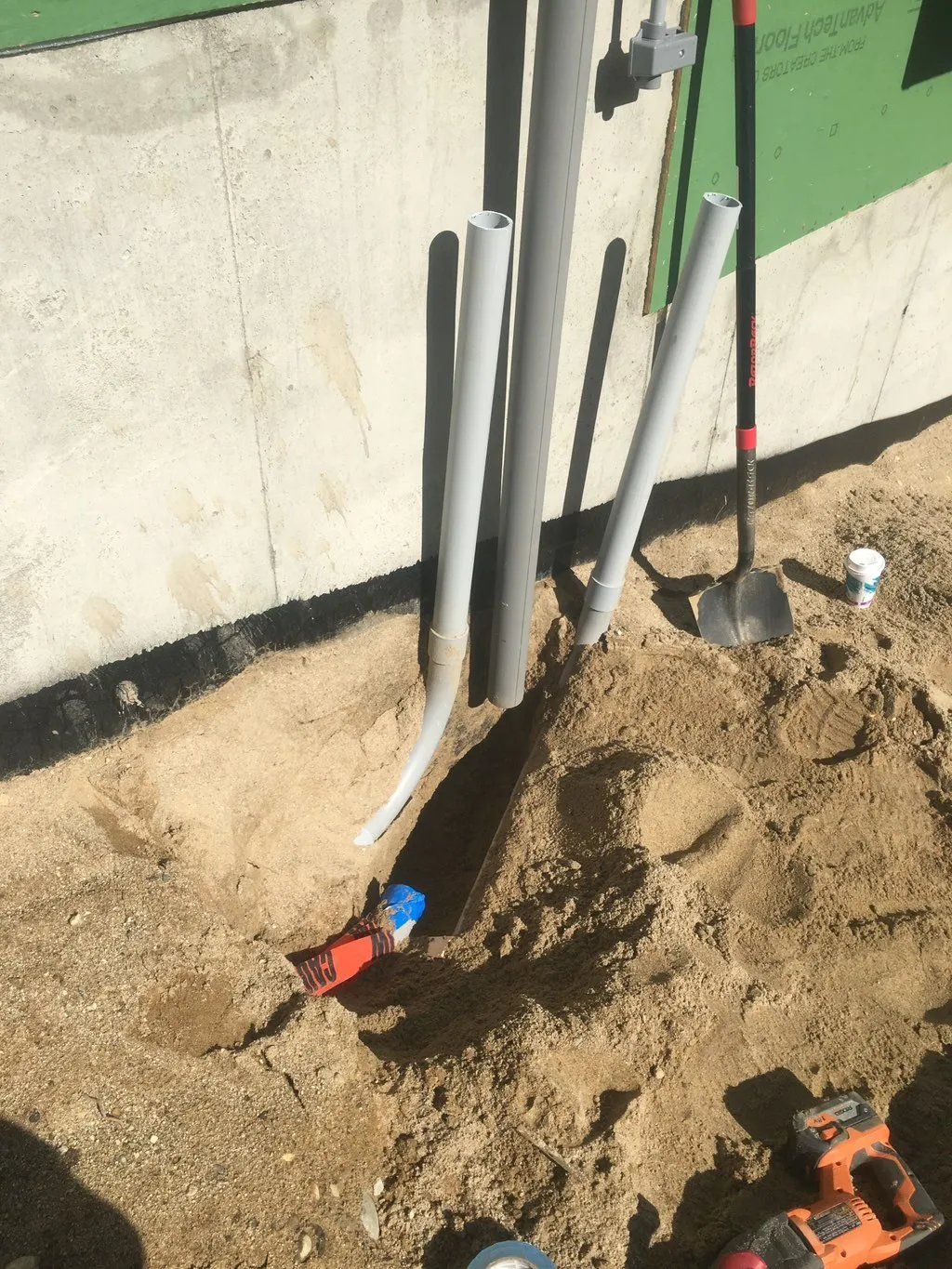 Electrical conduits emerging from the ground next to a concrete foundation. A shovel rests nearby.