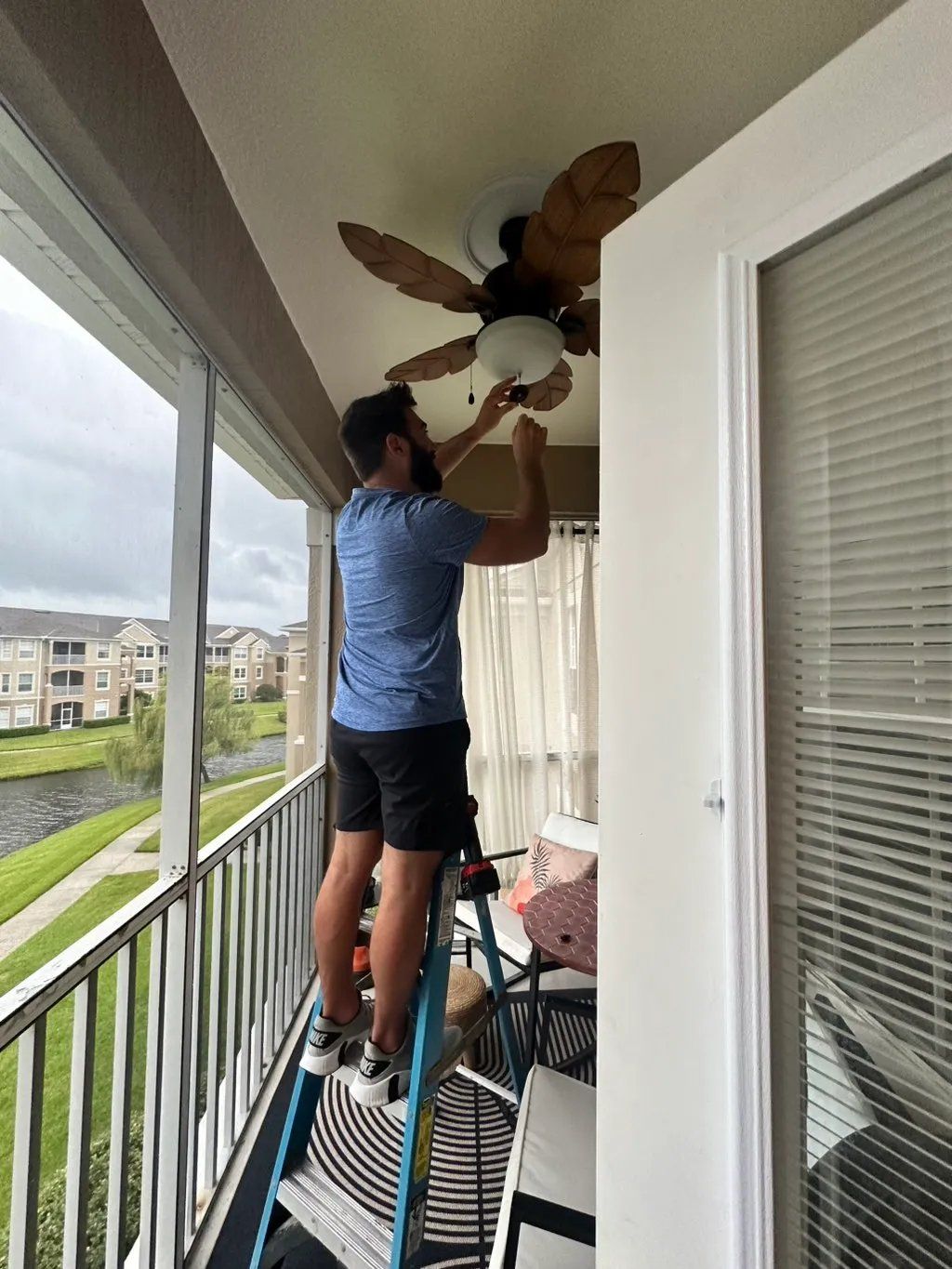 Man on ladder installing a light bulb in a ceiling fan on a balcony, overlooking a canal.