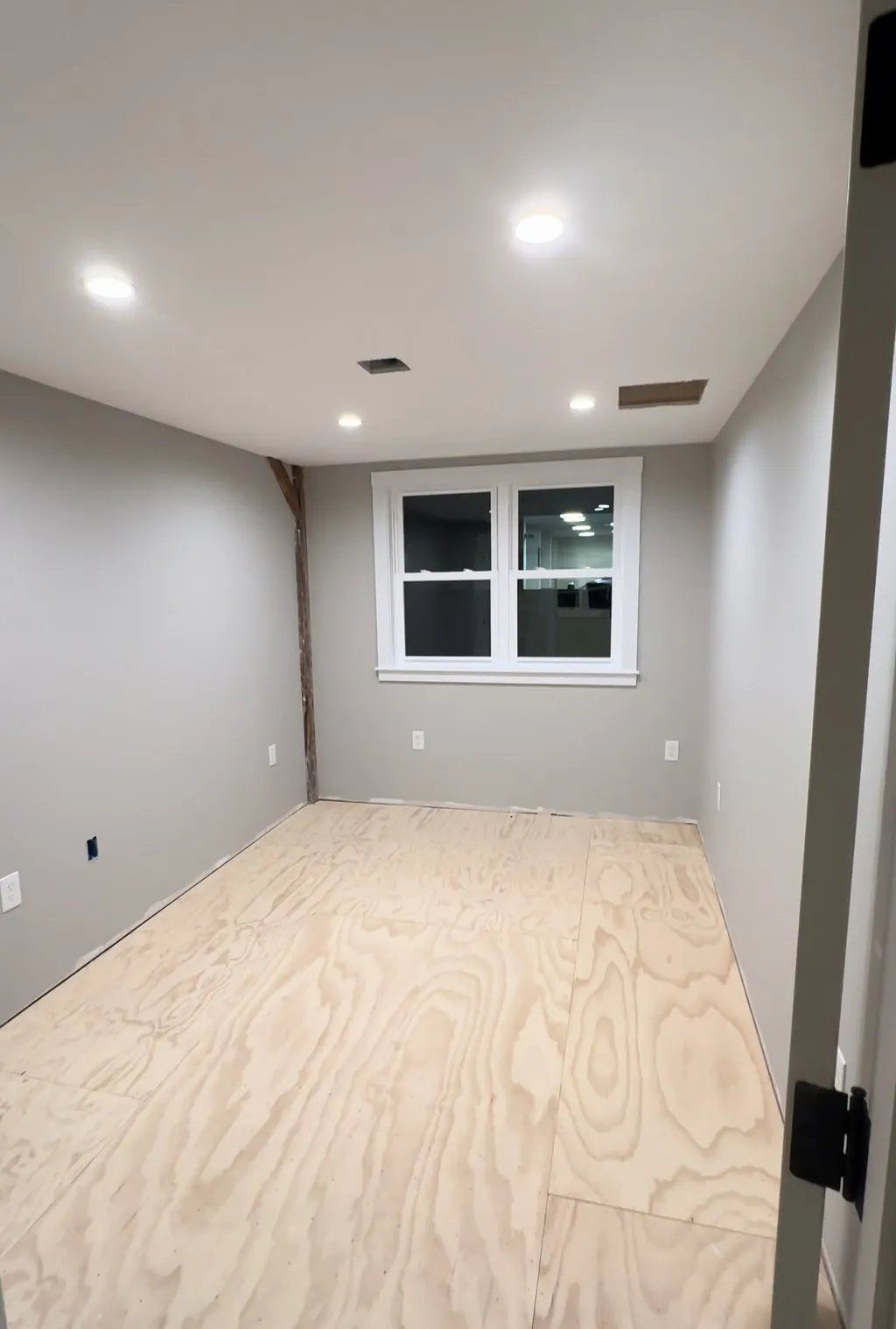 Empty room with plywood floor, gray walls, white window and trim, and recessed lights.