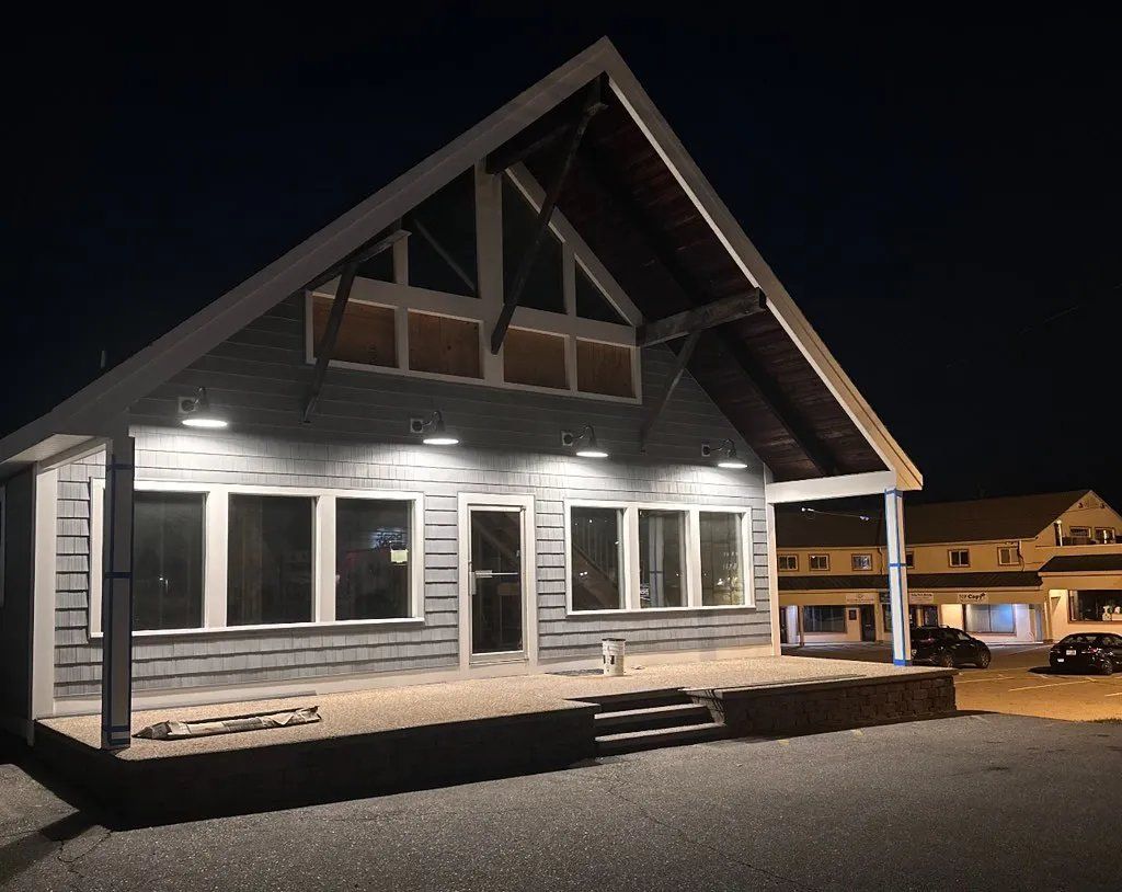 A gray building at night with lit windows, porch, and an A-frame roof; gravel area.