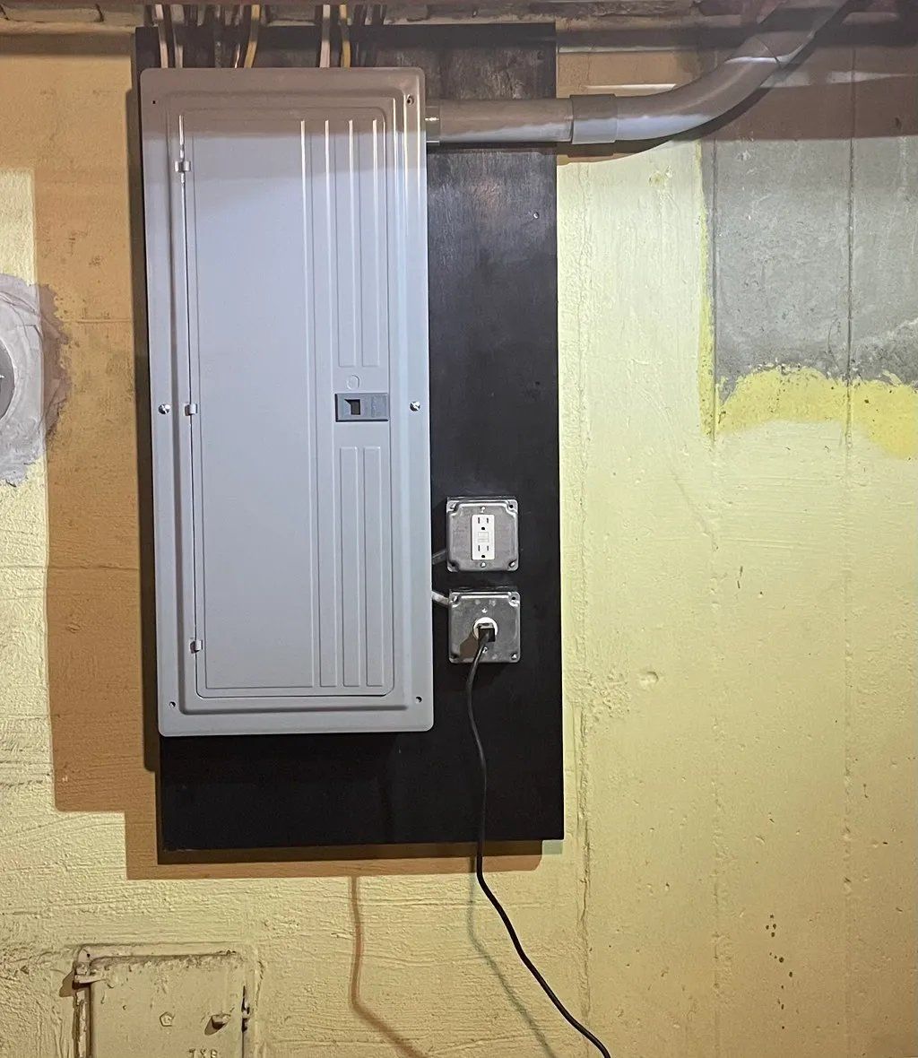 Electrical panel mounted on a black board on a yellow wall, with outlets and conduit.