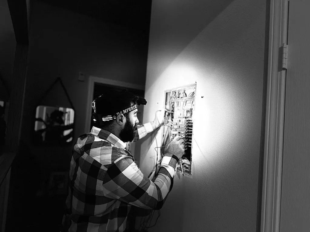 Man in plaid shirt examines a document on a wall. Bright light illuminates the wall and him.