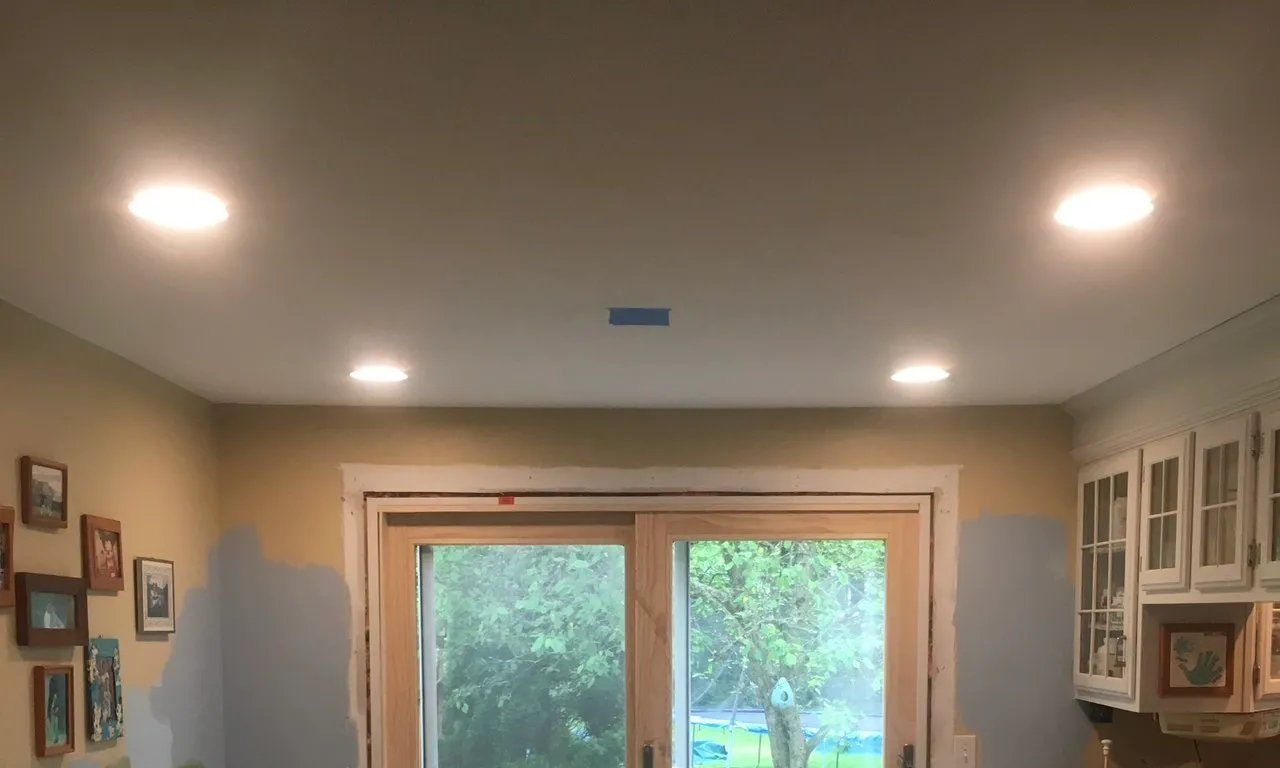 Recessed ceiling lights in a room with a sliding glass door and kitchen cabinets.