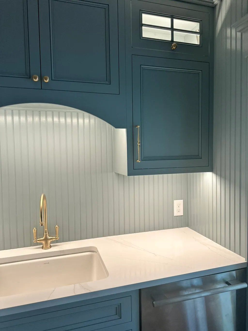 Blue kitchen cabinets with white countertop and sink, gold faucet.