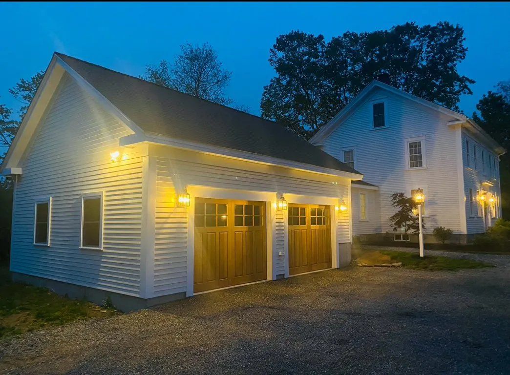 White two-car garage with wood doors and exterior lights, next to a two-story house, at dusk.