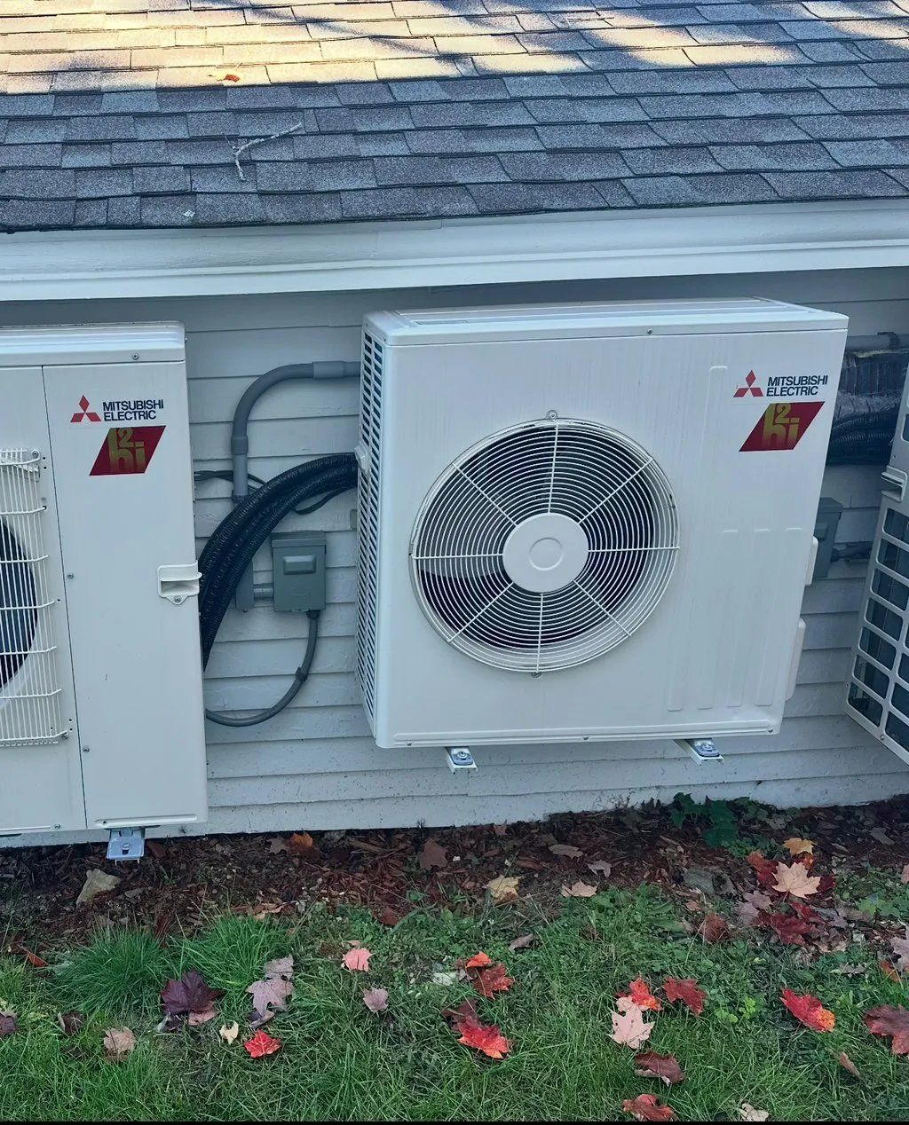 Three white Mitsubishi outdoor air conditioning units mounted on a white wall, with a gray roof above 