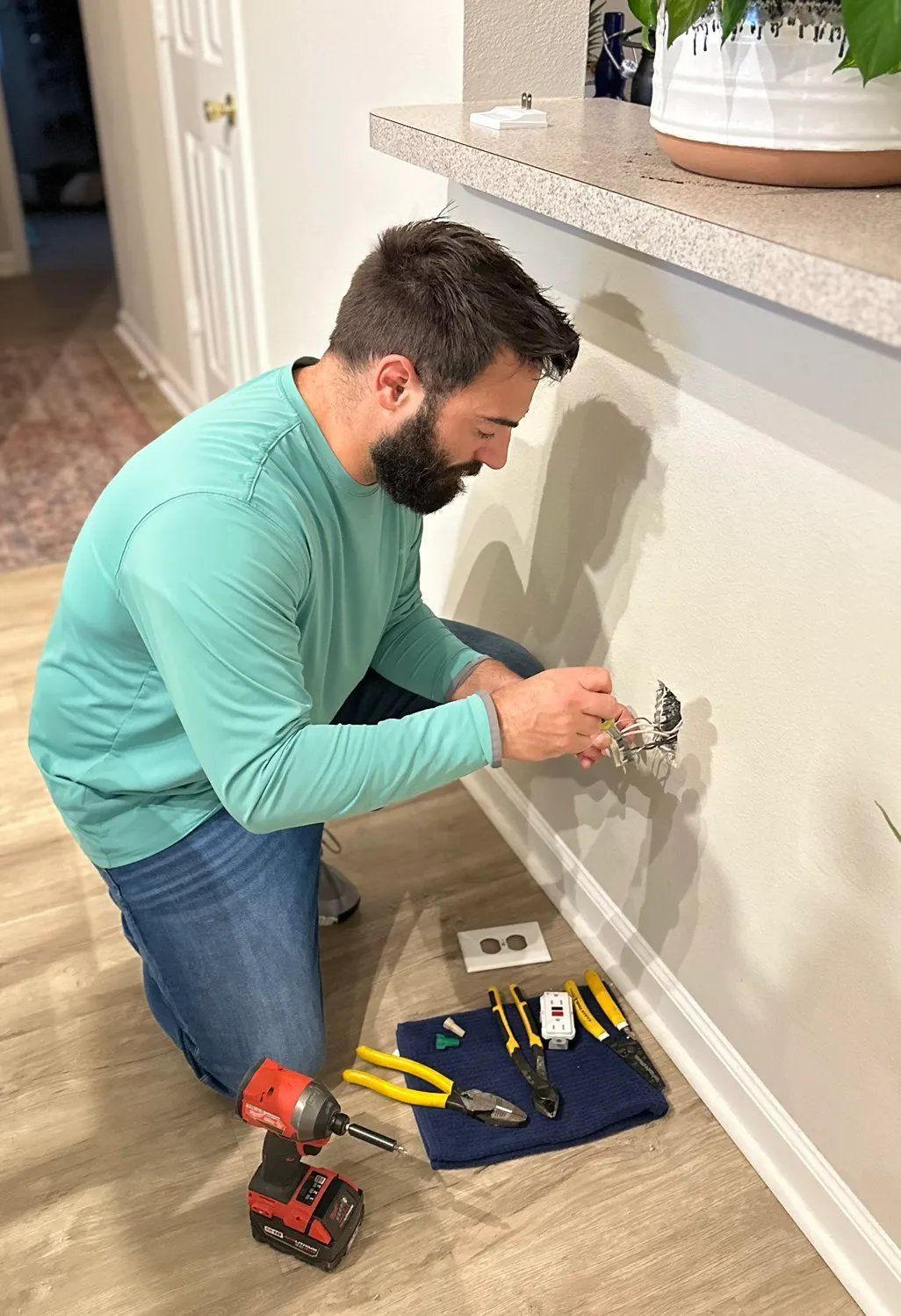 Man in teal shirt, jeans, installs electrical outlet on wall, tools on floor.