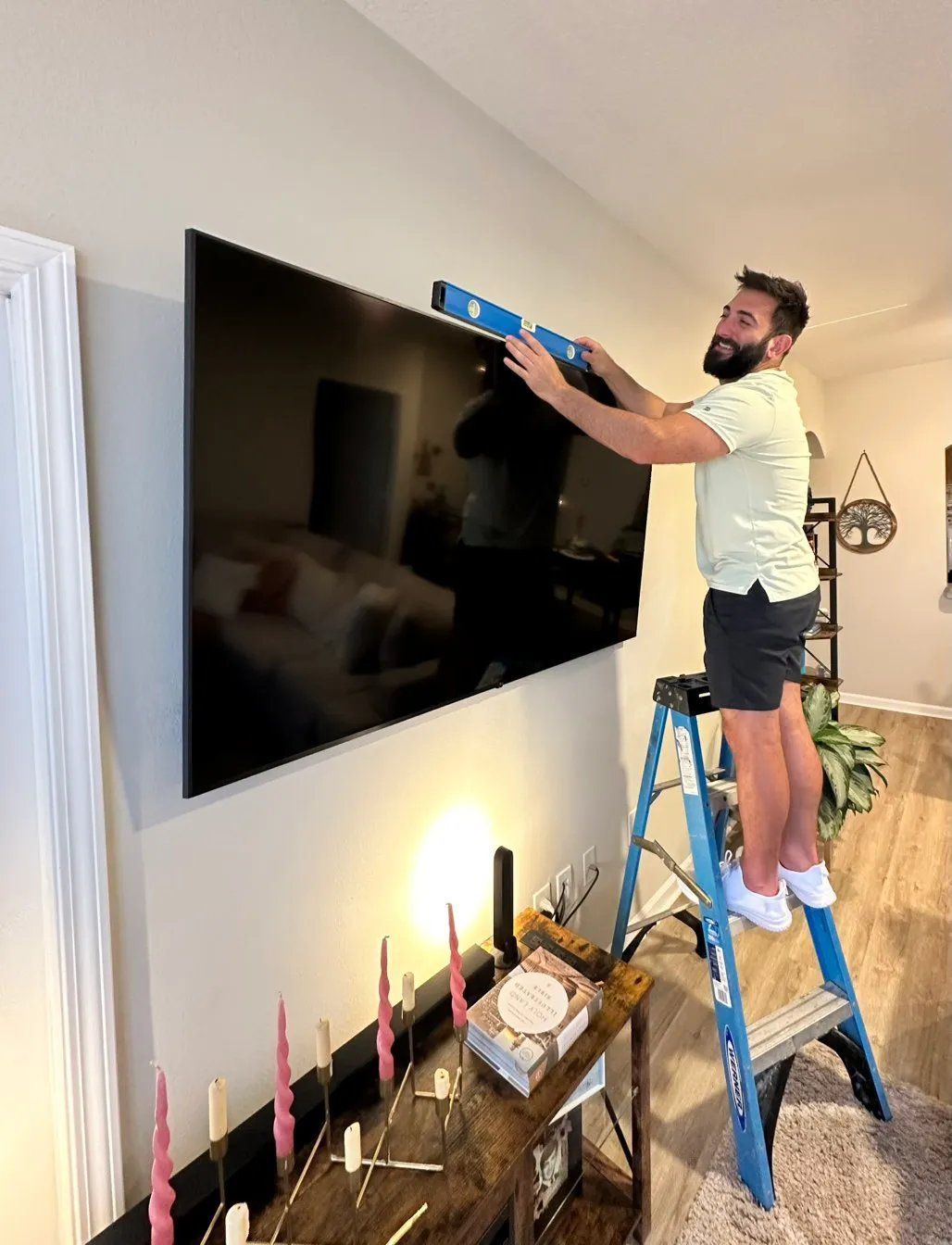 Man on ladder using level to hang a large TV on a beige wall in a home.