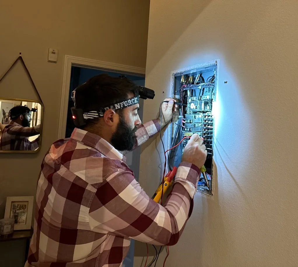 Electrician with headlamp working on an electrical panel, indoors, wearing a plaid shirt.