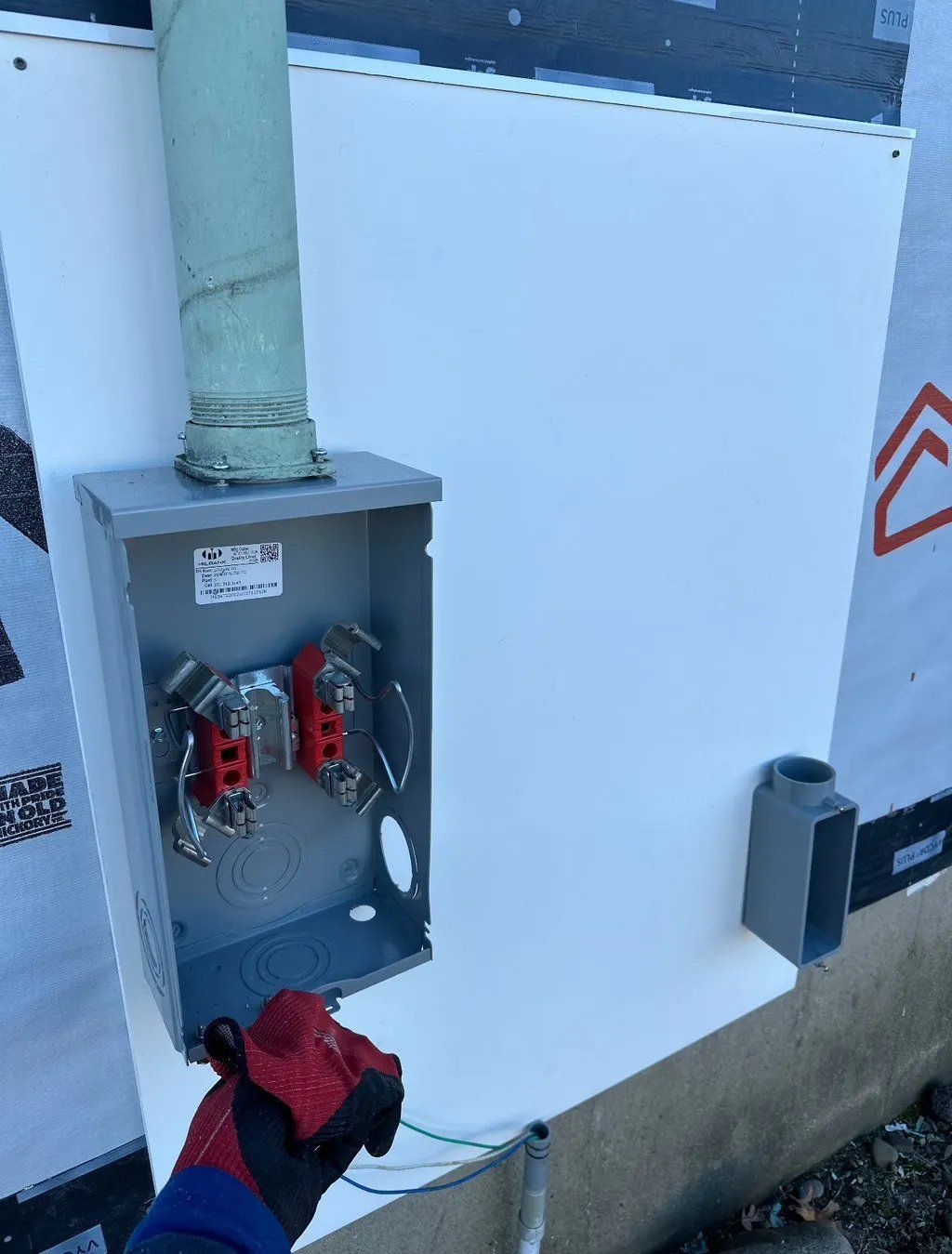 Exterior electrical box on a white wall, with a gloved hand holding a wire. Conduit pipe is visible.