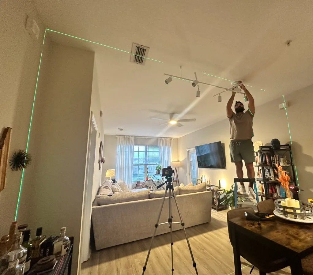 A person on a ladder installs a light fixture. A green laser line shows alignment on a ceiling.