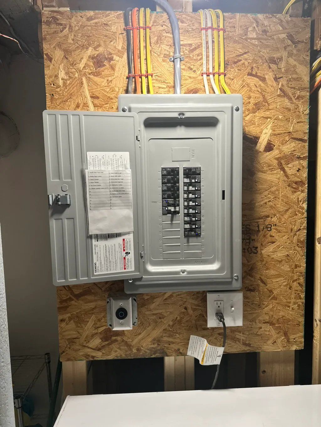 Electrical panel with open door, mounted on a wood surface, with wires and outlets nearby.
