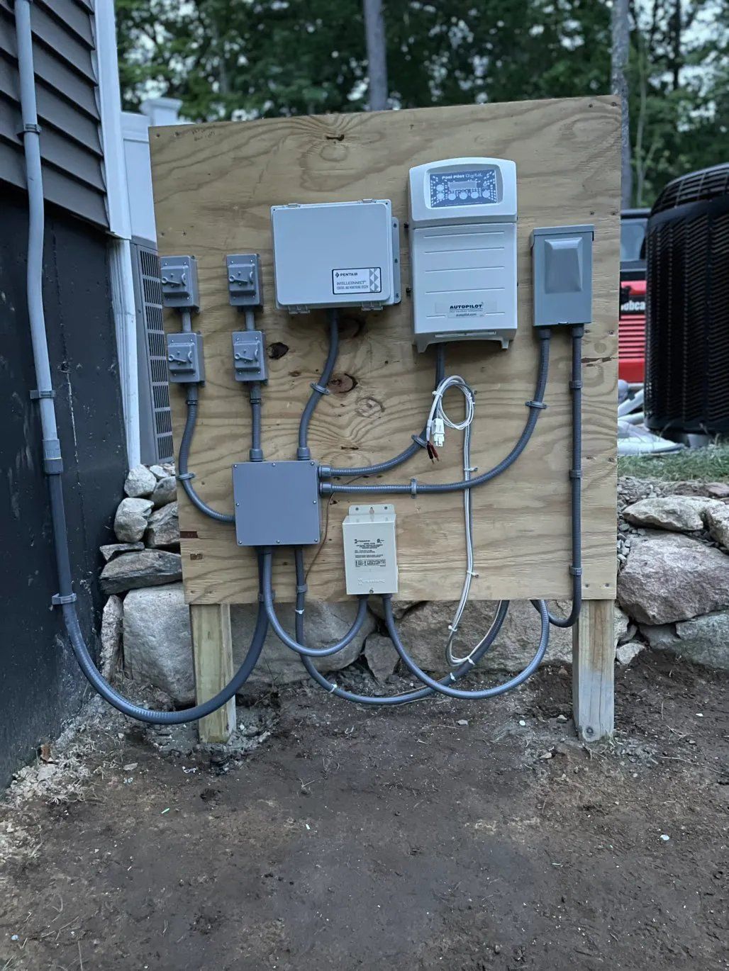 Electrical components mounted on a wooden board outdoors. Wires, boxes, and a control panel are visible.