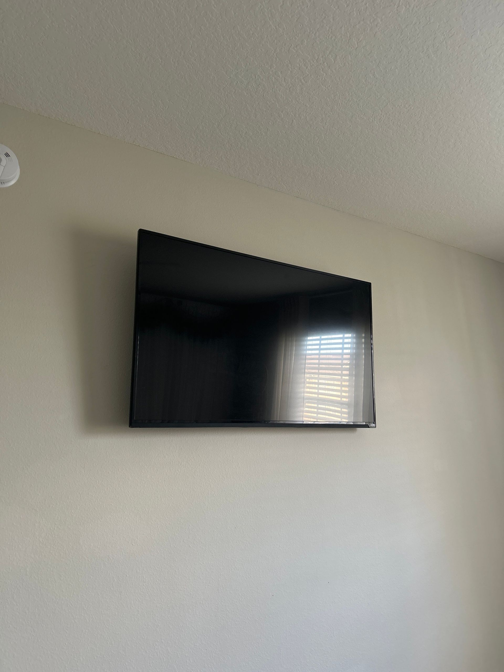 A wall-mounted black TV on a beige wall, reflecting window light.