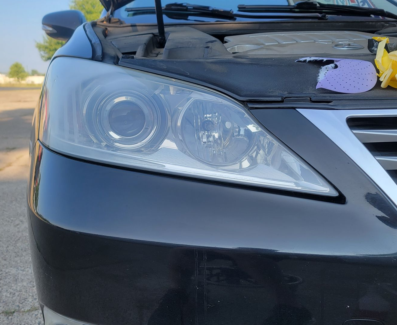 Black Lexus car front with foggy headlights, parked outdoors near a road.
