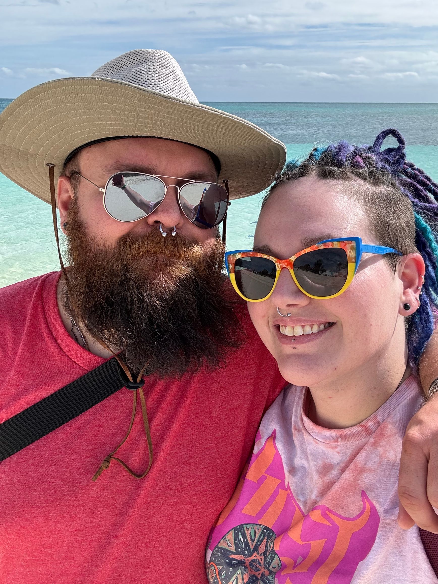 A man with a beard and a woman wearing sunglasses are posing for a picture on the beach.