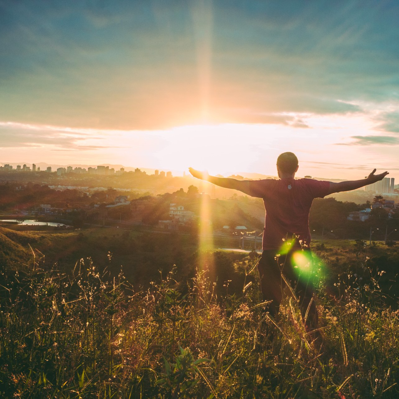Person with arms outstretched, facing sunset over city.