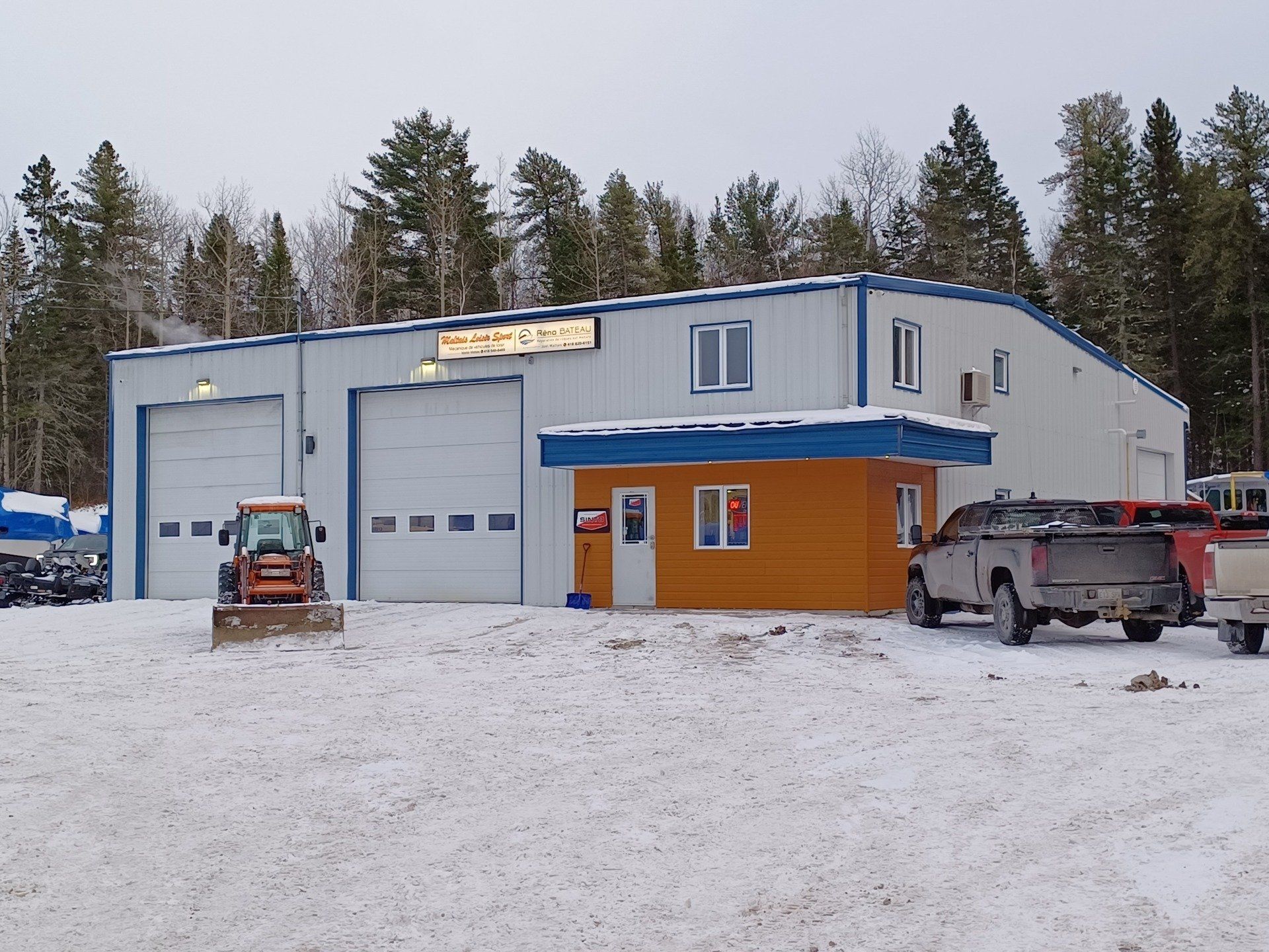 Bâtiment métallique avec portes de garage, petit bureau, neige, véhicules.