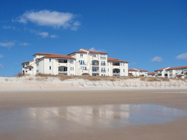 A large white building with a red roof sits on top of a sandy beach.