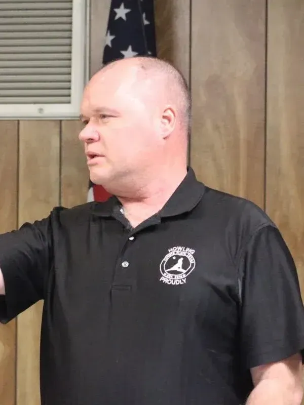 A man in a black shirt is standing in front of an american flag