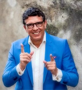 Ein Mann in einem blauen Anzug und mit Brille lächelt und zeigt in die Kamera.