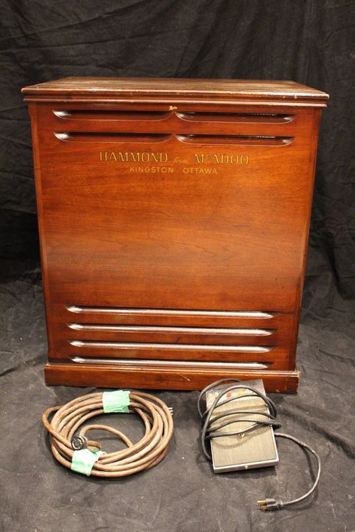 Wooden Hammond organ speaker cabinet with power cord and foot pedal on a black background.