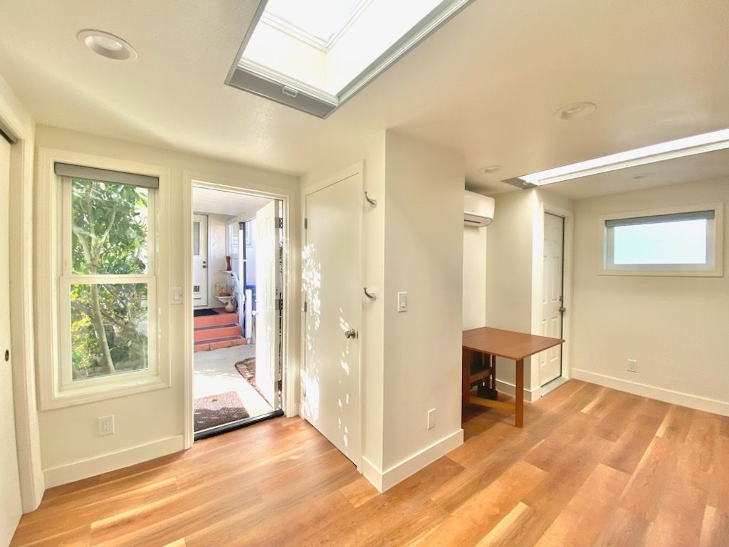 Bright interior room with wooden floors, white walls, and a table. Open doorway leads to another room.