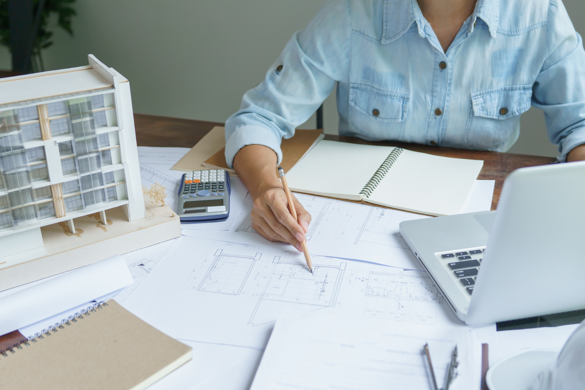 Architect at a desk, working on blueprints, with a building model and laptop.