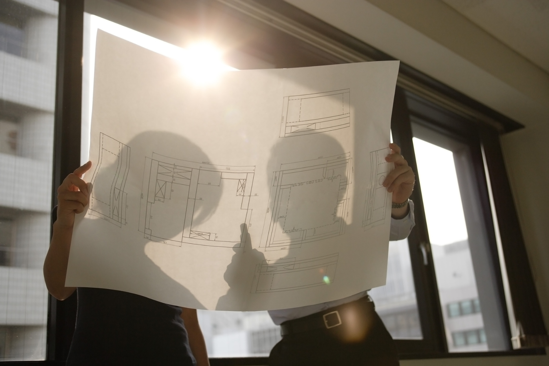 Two people looking at a blueprint, backlit by the sun in an office.