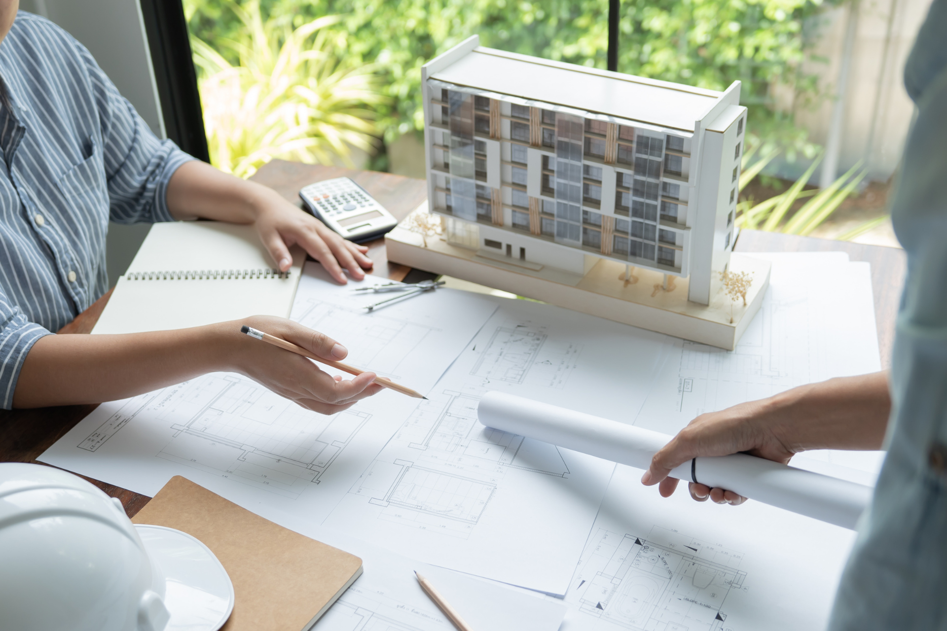 Two people reviewing architectural plans at a table with a building model and hard hats.