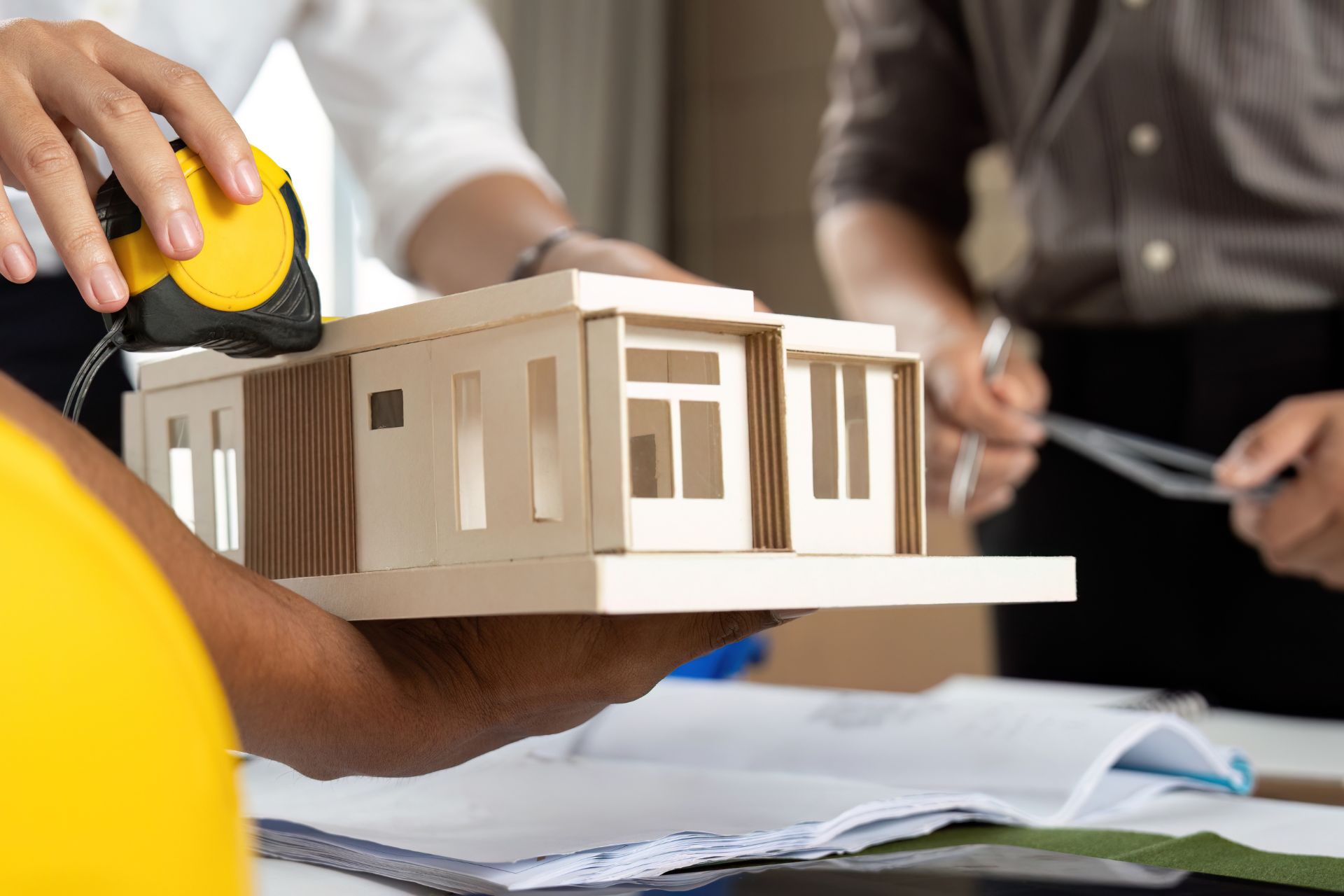 Construction workers review a model house with measuring tape and blueprints.