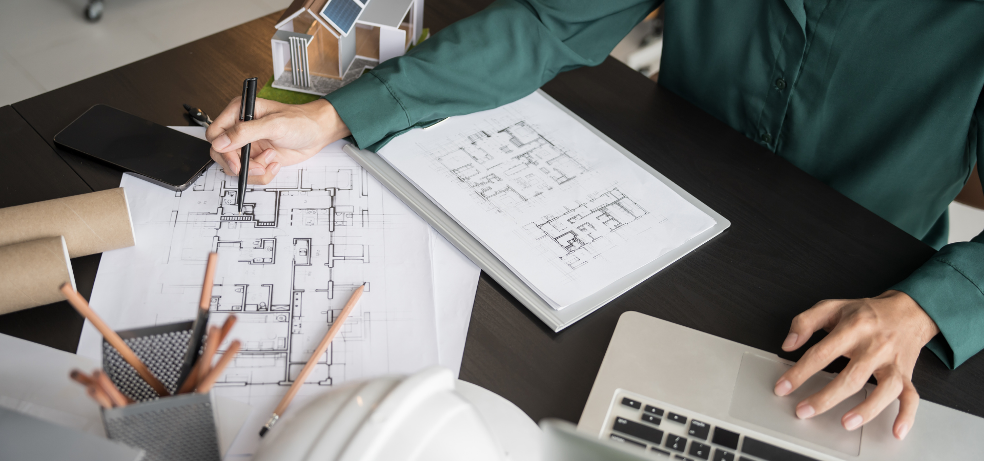 Person sketching a floor plan at a desk with papers, pencils, and a laptop open to blueprints