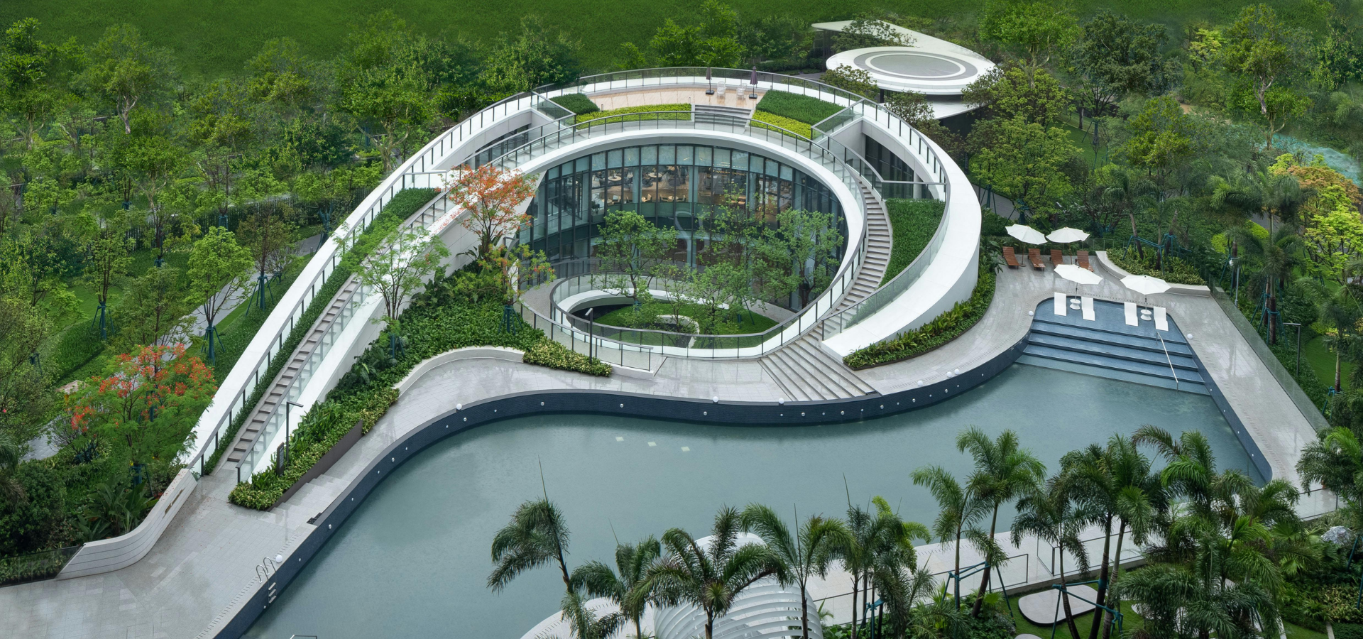 Aerial view of a modern curved building complex beside a pool, surrounded by lush greenery.