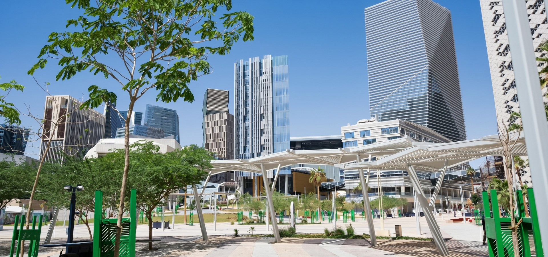 Modern city plaza with tall glass skyscrapers, trees, and green benches under a bright sky