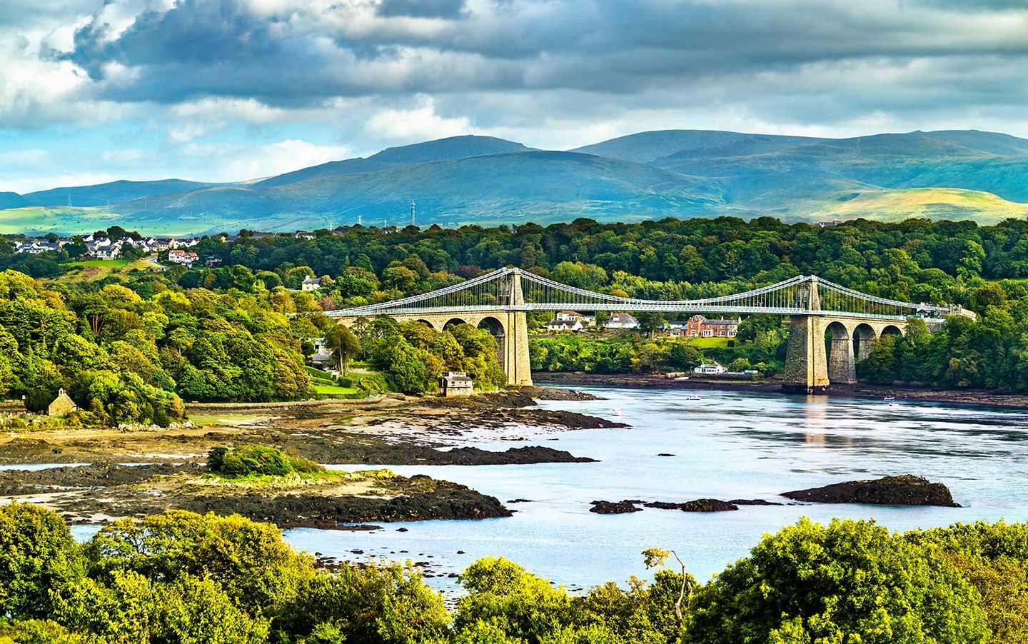 Bridge over water with mountains in background. Lush greenery and cloudy sky.