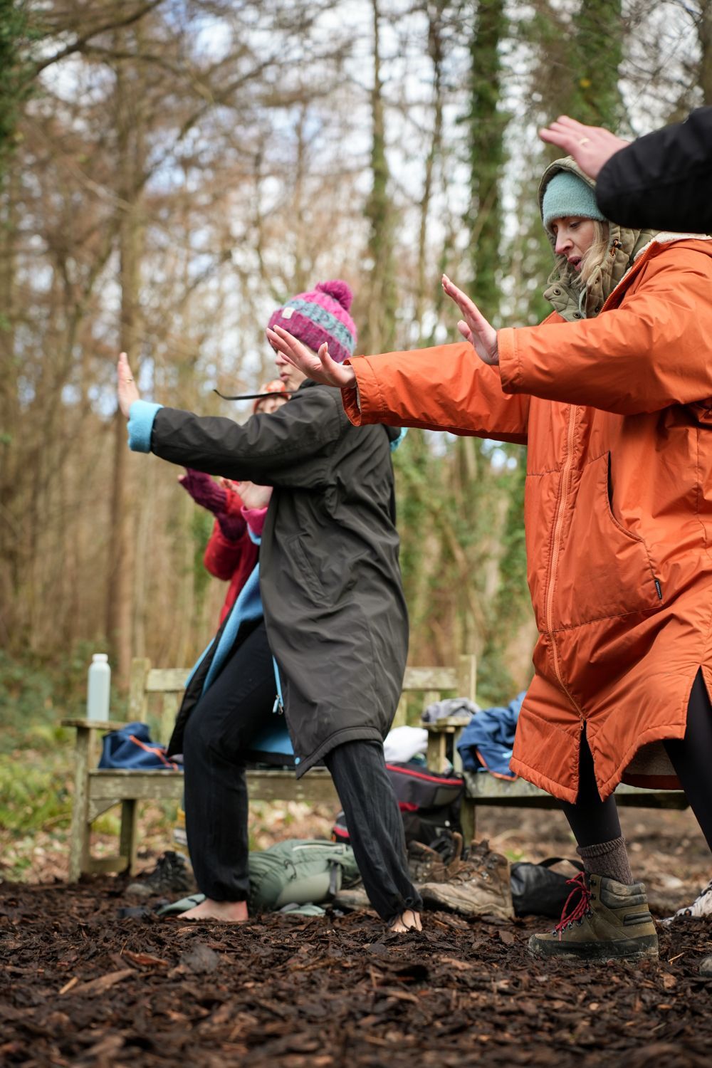 People in swimwear standing in a river, surrounded by trees. One woman laughs, others appear to be cold.