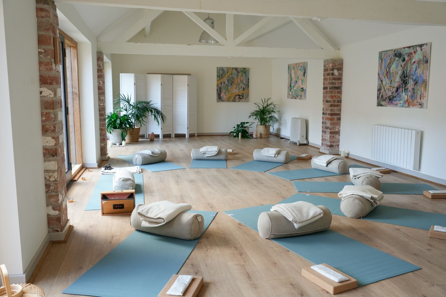 A light, airy yoga studio featuring blue mats arranged in a circle, each with a bolster and blanket on a wood floor.