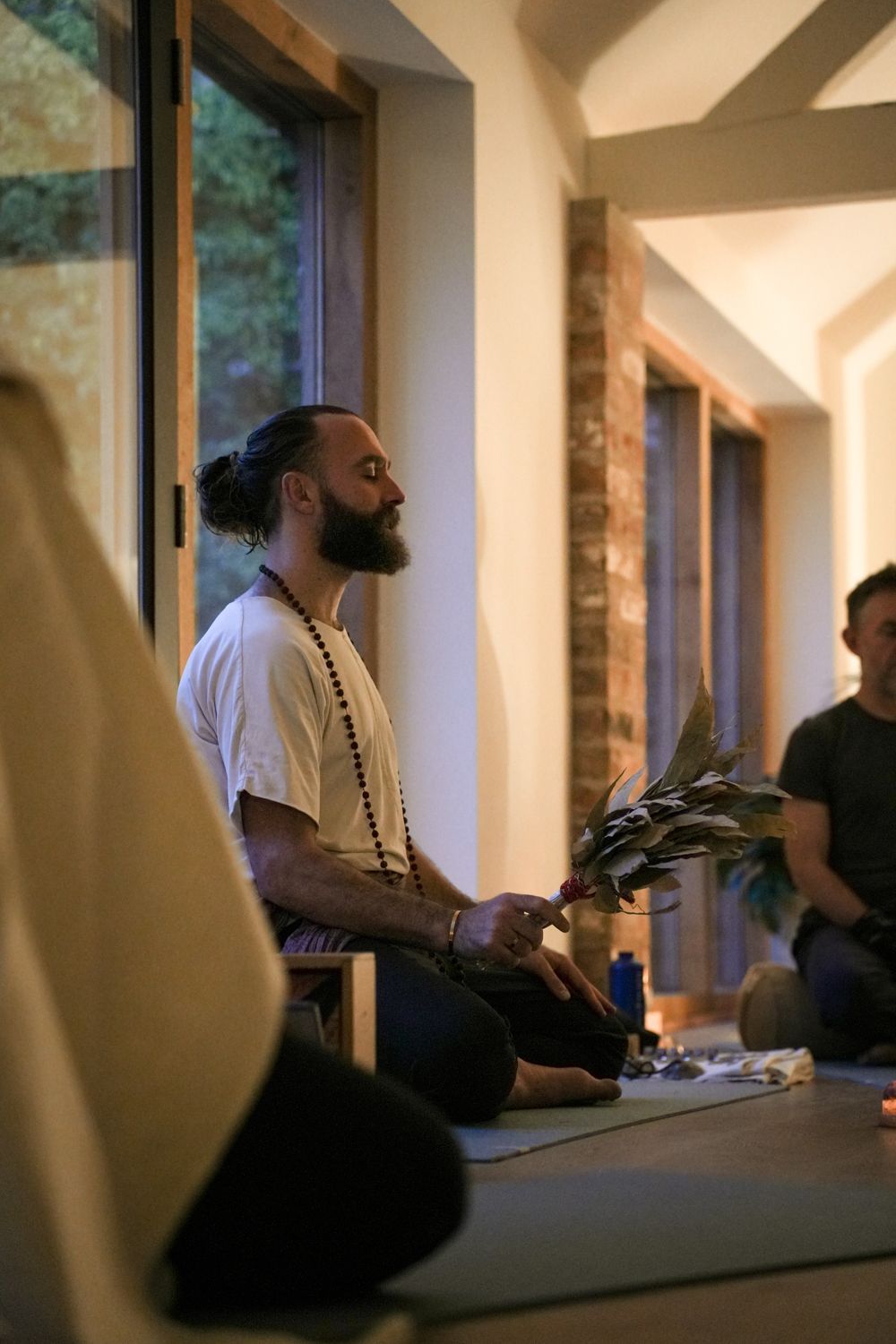 A person with a beard and hair in a bun sitting in a meditative pose while holding a leafy bundle in a bright room.