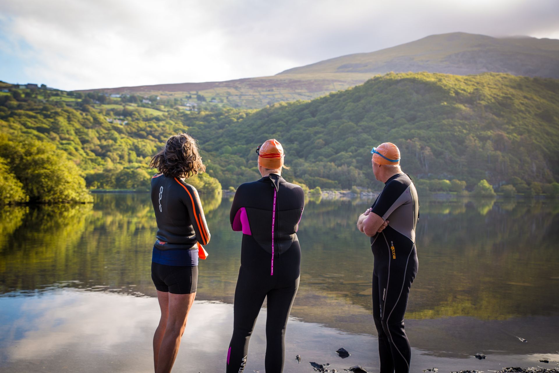 Three people in wet suits are standing on the shore of a lake.