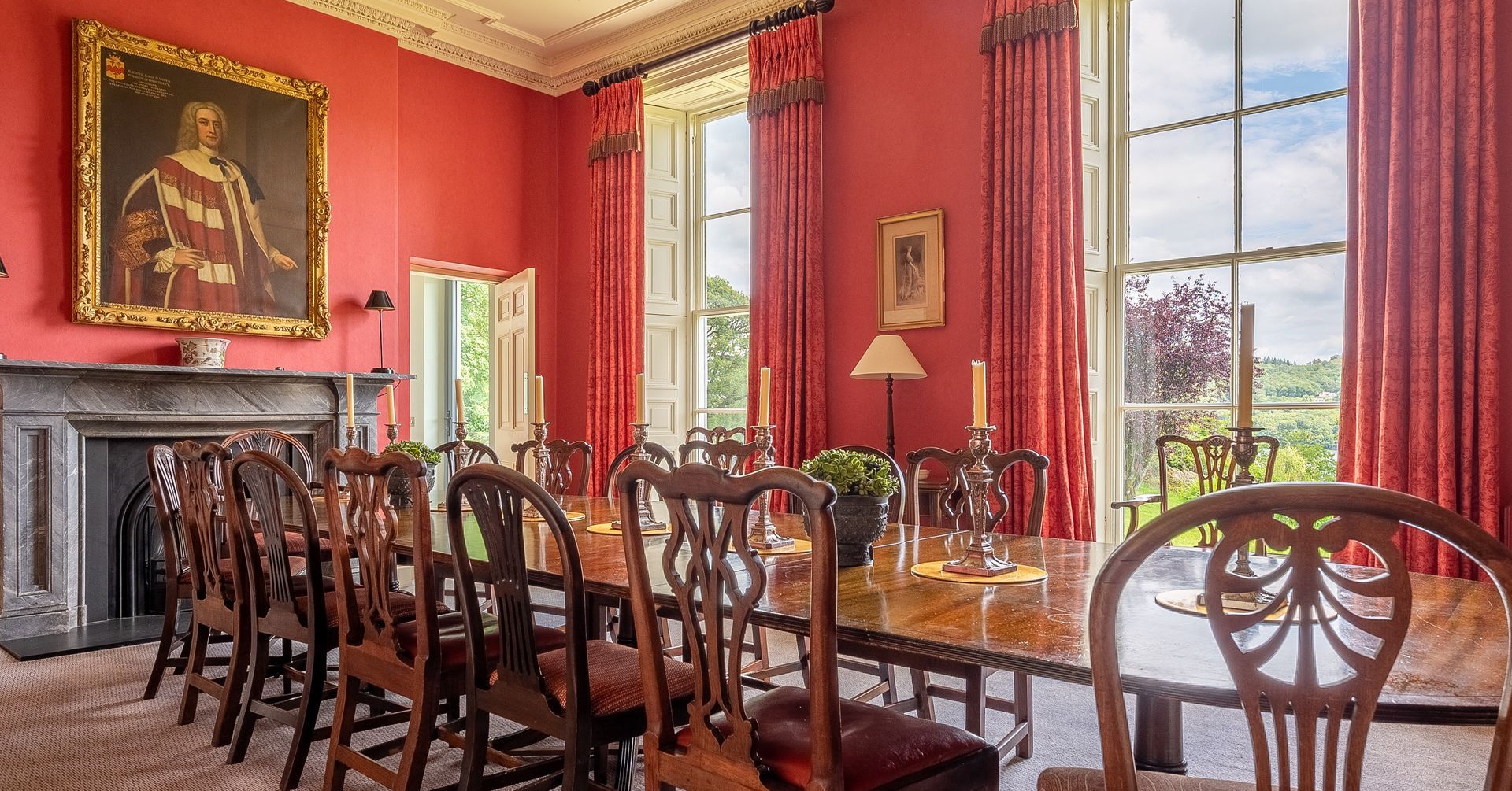 Formal dining room with red walls, long wooden table, ornate chairs, large windows, and a portrait.