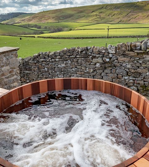 Wooden hot tub with bubbly water, set against a stone wall and rolling green hills.