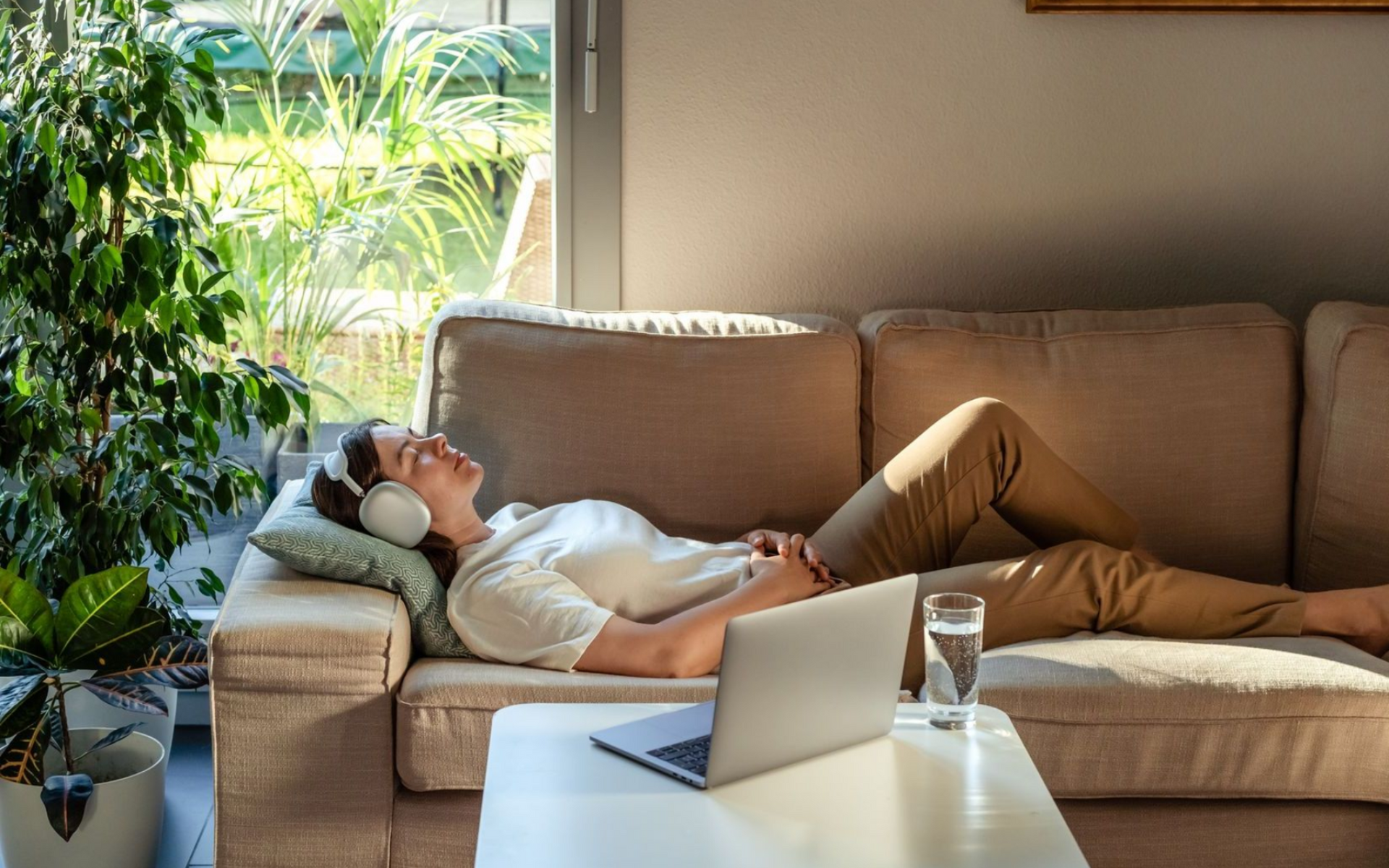 Woman relaxing on a couch, wearing headphones, eyes closed. Laptop and water glass on a table. Sunlight streams in.
