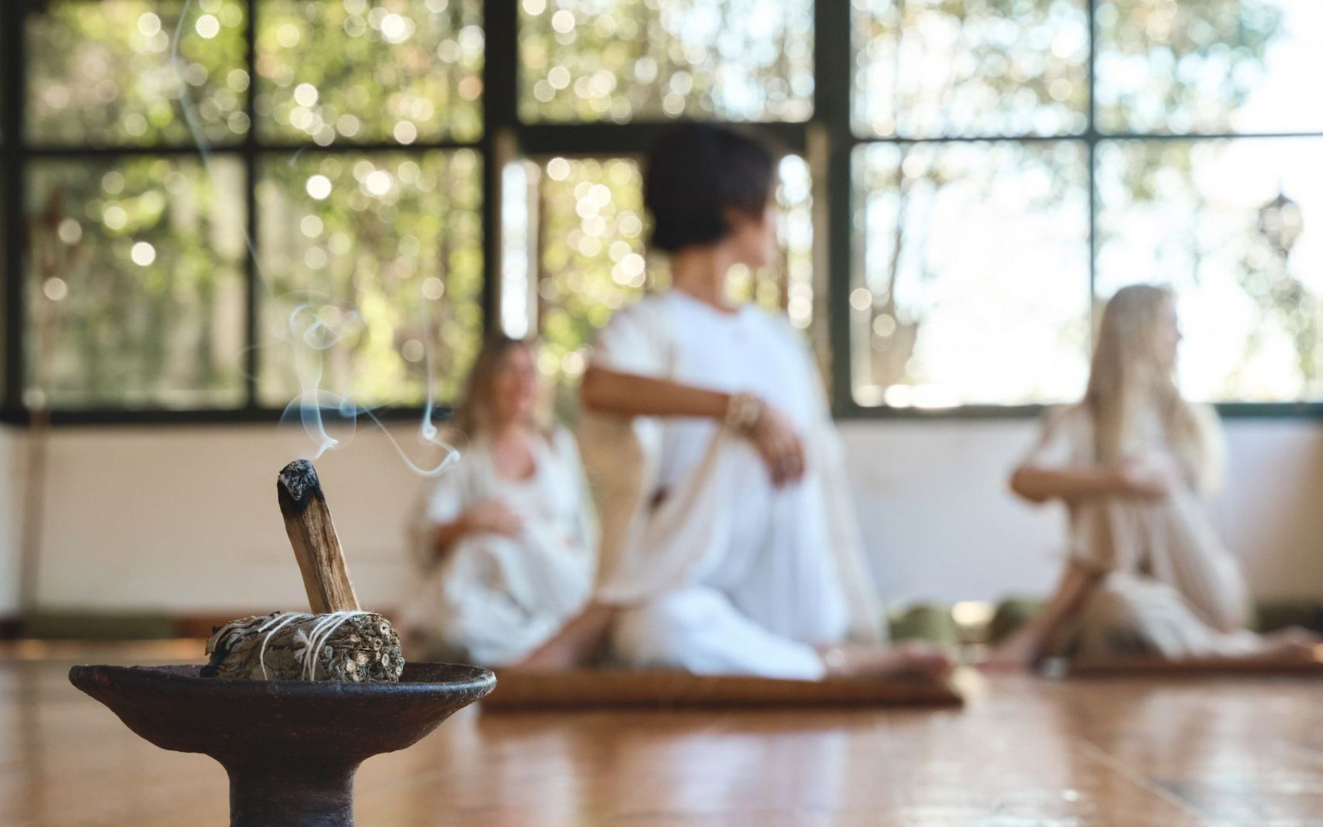 Incense burning, three people meditating in light-filled room.