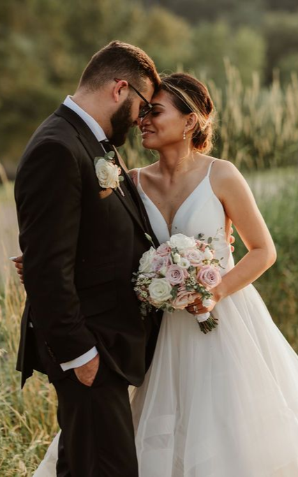 A bride and groom standing closely together, holding a bouquet, forehead to forehead in a grassy outdoor setting.