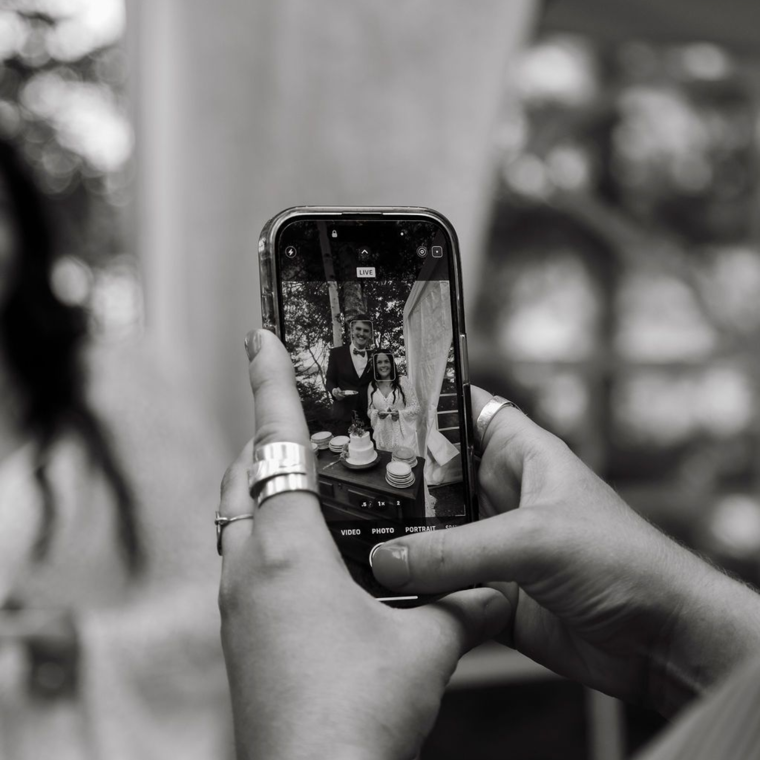 Close-up of hands holding a smartphone, capturing a photo of a couple posing by a small wedding cake.